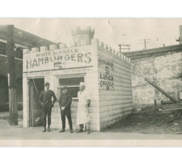 Three men stand outside a White Castle location in 1921. The building looks like a castle made of white bricks with a tower coming from the center of the roof.
