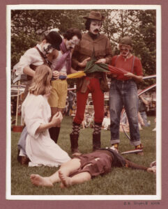 Four mimes stand in a line, one kneels on the ground, and one lies face-up in the grass.