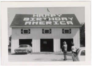 Two people looking up at a man seated on the roof of a large garage, where "Happy Birthday America" has been written.